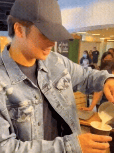 A Man Wearing A Hat And A Denim Jacket Is Pouring Milk Into A Cup . GIF
