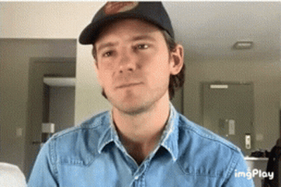 A Man Wearing A Hat And A Denim Shirt Looks At The Camera GIF