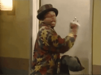 A Man Wearing A Hat And A Floral Shirt Is Standing In Front Of A Door And Giving The Middle Finger . GIF