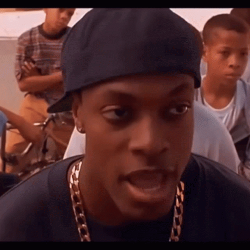 A Man Wearing A Hat And A Gold Chain Is Talking To A Group Of People . GIF