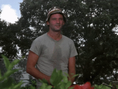 A Man Wearing A Hat And A Gray Shirt Is Standing In A Garden GIF