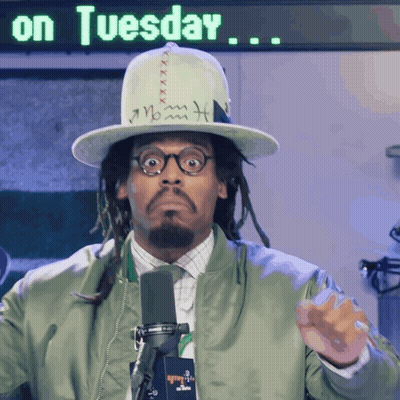 A Man Wearing A Hat And A Green Jacket Stands In Front Of A Sign That Says " On Tuesday " GIF