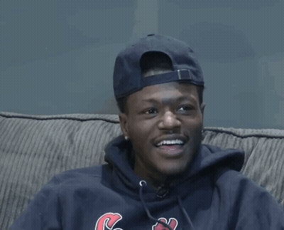 A Man Wearing A Hat And A Hoodie With The Letter C On It Sits On A Couch GIF