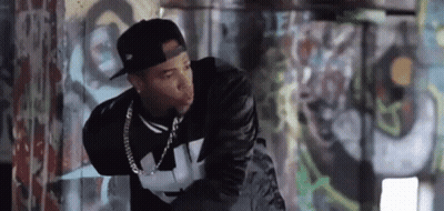 A Man Wearing A Hat And A Leather Jacket Is Standing In Front Of A Graffiti Wall . GIF