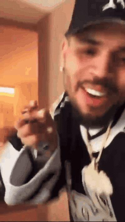 A Man Wearing A Hat And A Necklace Is Holding A Pair Of Shoes . GIF