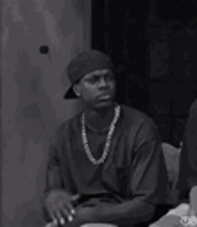 A Man Wearing A Hat And A Necklace Is Sitting At A Table With His Hands Folded . GIF