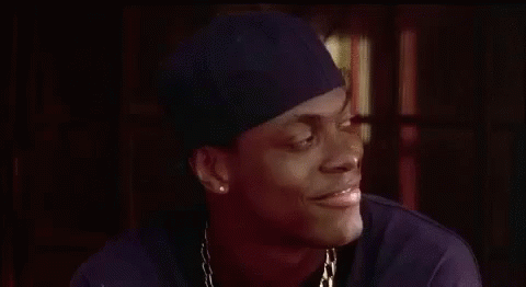 A Man Wearing A Hat And A Necklace Is Smiling And Looking At The Camera . GIF
