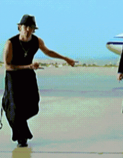 A Man Wearing A Hat And A Necklace Is Standing In Front Of An Airplane GIF