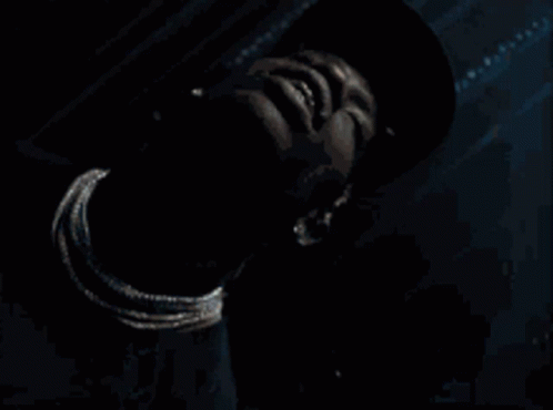 A Man Wearing A Hat And A Necklace Looks Down At Something GIF