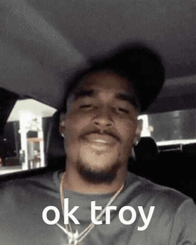 A Man Wearing A Hat And A Necklace Says Ok Troy On His Face GIF