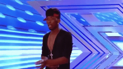 A Man Wearing A Hat And A Necklace Stands On A Stage GIF