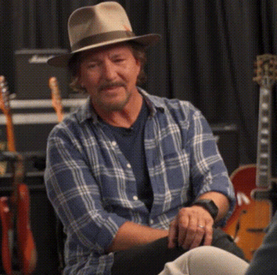 A Man Wearing A Hat And A Plaid Shirt Is Sitting In Front Of Guitars GIF