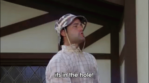 A Man Wearing A Hat And A Plaid Shirt Is Standing In Front Of A Building And Saying `` Its In The Hole '' . GIF