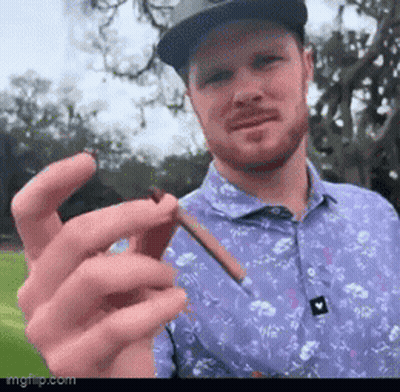 A Man Wearing A Hat And A Purple Shirt Is Holding A Cigarette In His Hand . GIF