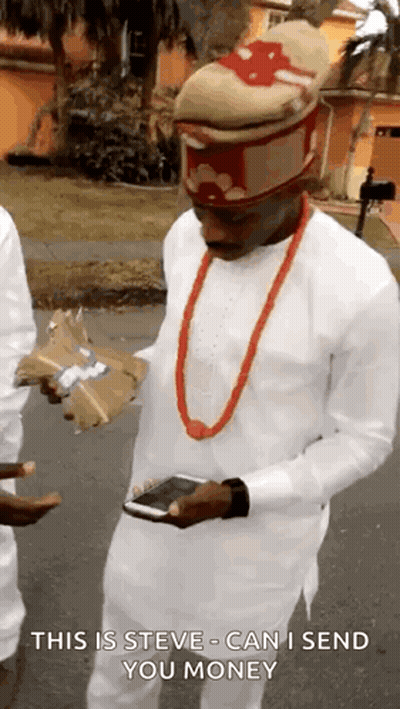 A Man Wearing A Hat And A Red Necklace Is Holding A Cell Phone And A Bag Of Money . GIF