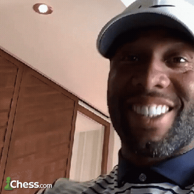 A Man Wearing A Hat And A Striped Shirt Is Smiling In Front Of A Sign That Says Chess.com GIF