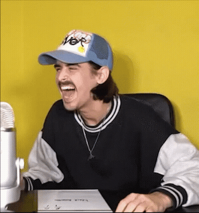 A Man Wearing A Hat And A Sweater Is Sitting At A Desk Laughing . GIF
