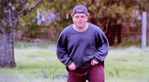 A Man Wearing A Hat And A Sweater Is Standing In A Grassy Field . GIF