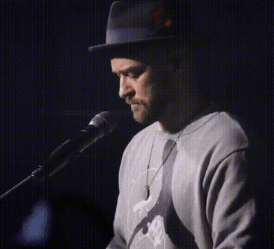 A Man Wearing A Hat And A Sweater Singing Into A Microphone GIF