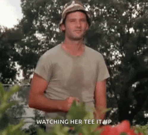 A Man Wearing A Hat And A T-Shirt Is Standing In Front Of A Garden . GIF