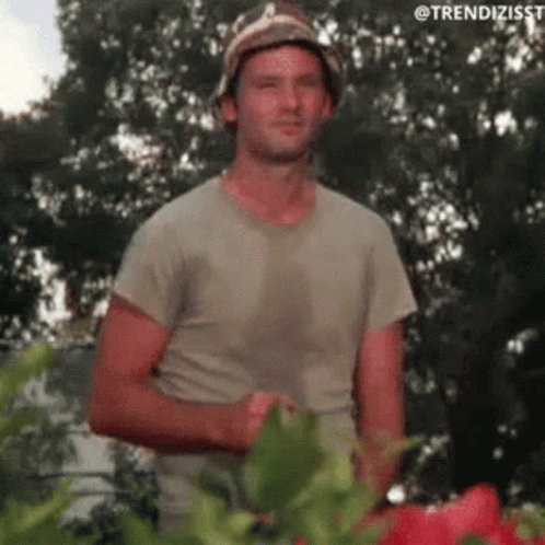 A Man Wearing A Hat And A T-Shirt Is Standing Next To A Plant . GIF
