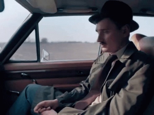 A Man Wearing A Hat And A Trench Coat Sits In The Back Seat Of A Car GIF