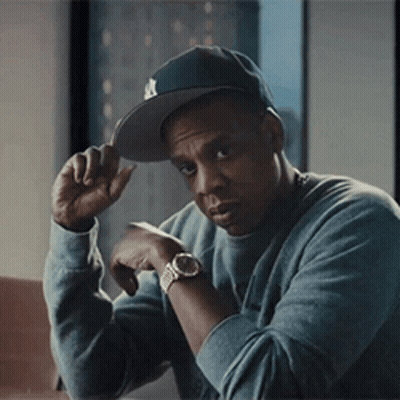 A Man Wearing A Hat And A Watch Is Sitting At A Table With His Hand On His Hat . GIF