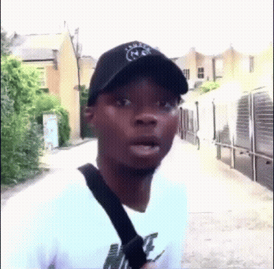 A Man Wearing A Hat And A White Shirt Is Standing On A Sidewalk . GIF