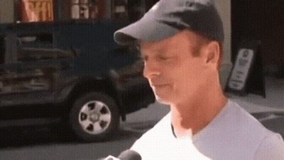 A Man Wearing A Hat And A White Shirt Is Talking Into A Microphone On The Street . GIF