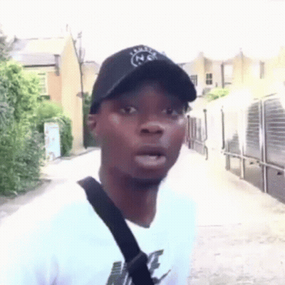 A Man Wearing A Hat And A White T-Shirt Is Standing On A Sidewalk . GIF