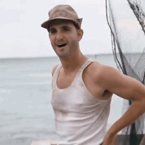 a man wearing a hat and a white tank top is standing on a boat in the ocean .
