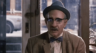 A Man Wearing A Hat And Bow Tie Is Sitting In Front Of A Door . GIF