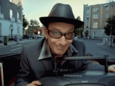 A Man Wearing A Hat And Glasses Is Driving Down A Street GIF