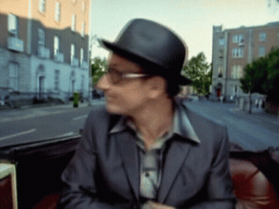 A Man Wearing A Hat And Glasses Is Sitting In Front Of A Blurry Street GIF