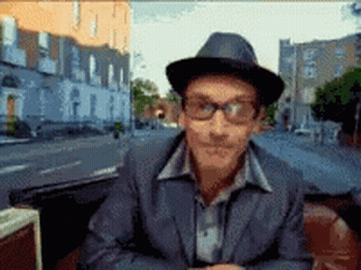 A Man Wearing A Hat And Glasses Is Sitting In Front Of A City Street GIF