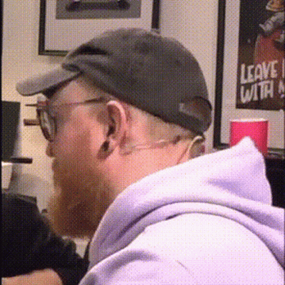 A Man Wearing A Hat And Glasses Is Sitting In Front Of A Leave With Poster GIF