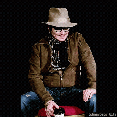 A Man Wearing A Hat And Glasses Is Sitting On A Chair With A Cup Of Coffee GIF