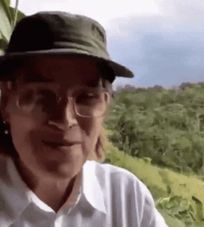 A Man Wearing A Hat And Glasses Is Standing In A Field . GIF