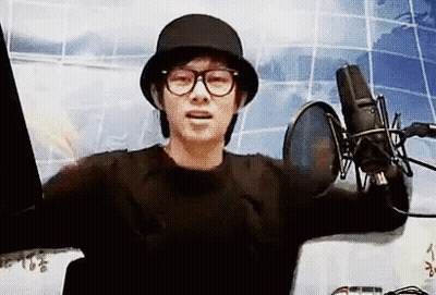A Man Wearing A Hat And Glasses Is Standing In Front Of A Microphone And Talking Into It . GIF