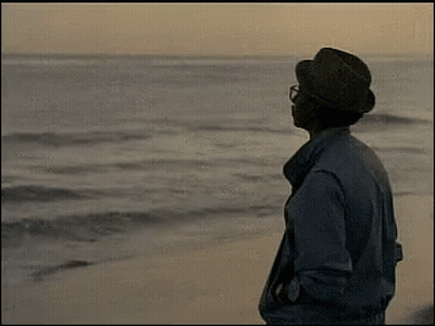 A Man Wearing A Hat And Glasses Is Standing On A Beach Looking At The Ocean GIF