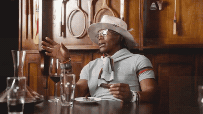 A Man Wearing A Hat And Glasses Sits At A Table With Wine Glasses And A Plate Of Food GIF