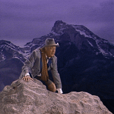 A Man Wearing A Hat And Scarf Is Sitting On A Rock In Front Of A Mountain GIF