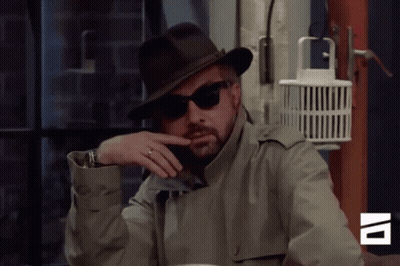 A Man Wearing A Hat And Sunglasses Is Sitting At A Table In Front Of A Sign That Says Av GIF