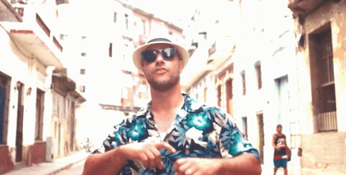 A Man Wearing A Hat And Sunglasses Is Standing In A Street GIF