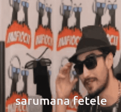 A Man Wearing A Hat And Sunglasses Is Standing In Front Of A Wall With The Words Sarumana Fetele Written On It . GIF
