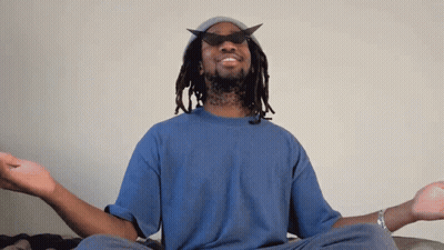 A Man Wearing A Hat And Sunglasses Sits In A Lotus Position With His Eyes Closed GIF