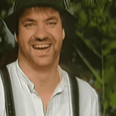 A Man Wearing A Hat And Suspenders Is Smiling And Looking At The Camera . GIF