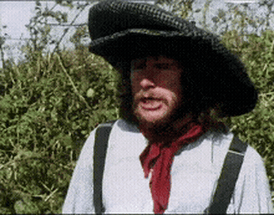 A Man Wearing A Hat And Suspenders Is Standing In Front Of A Bush GIF
