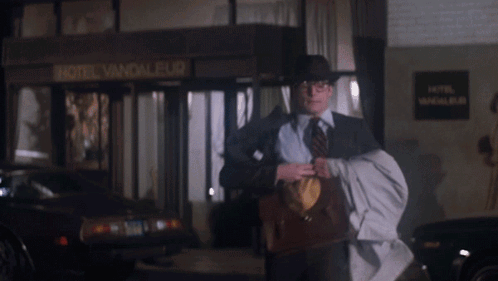 A Man Wearing A Hat And Tie Is Holding A Briefcase With The Letter S On The Front GIF