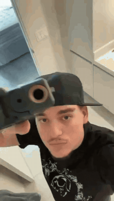 A Man Wearing A Hat Is Holding A Gun To His Face . GIF